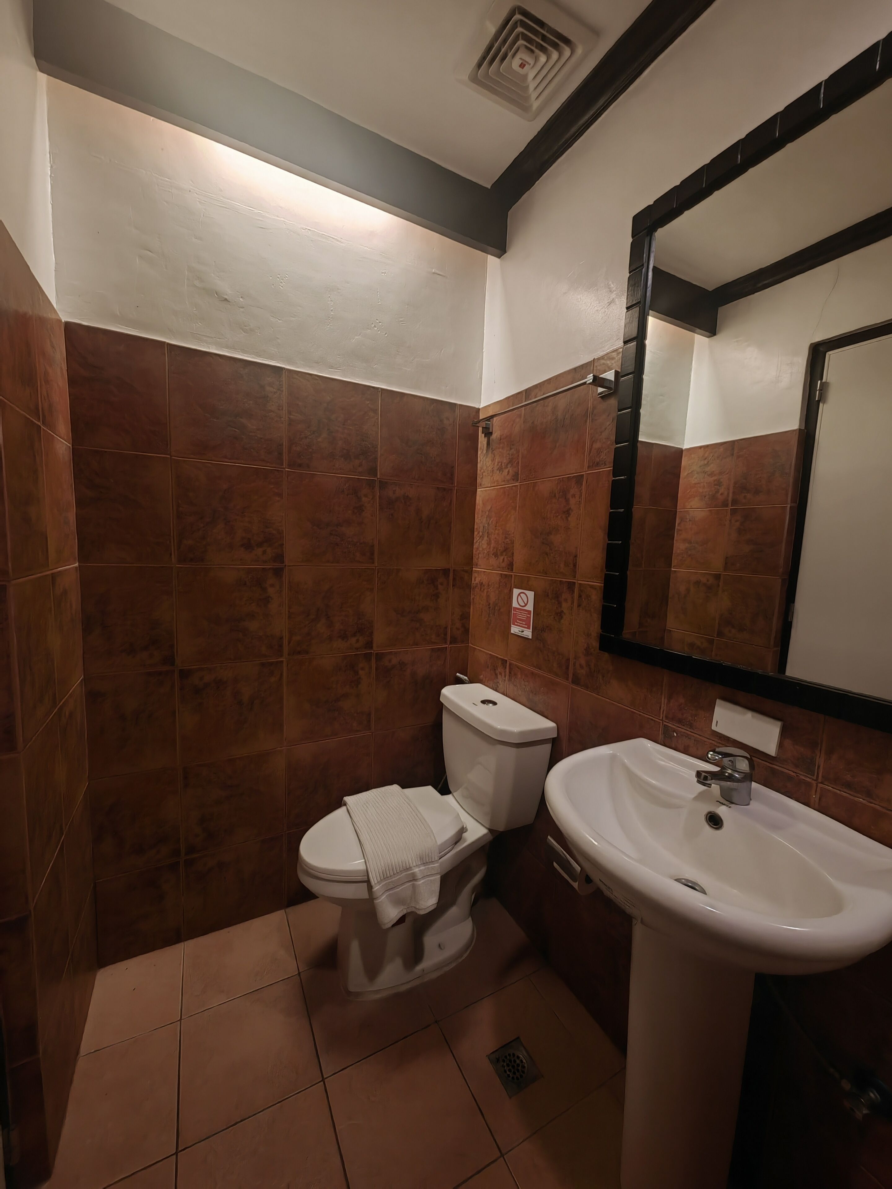 A bathroom with brown tiles, a white toilet, a white sink, and a mirror.