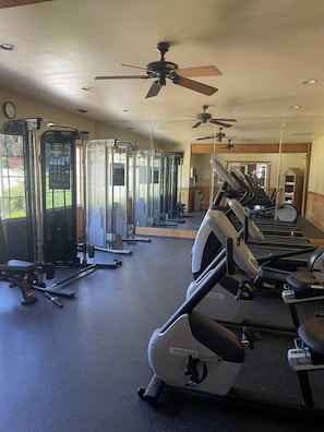 Un gimnasio con diverso equipo de ejercicio, incluyendo cintas de correr y máquinas de pesas, y ventiladores de techo.