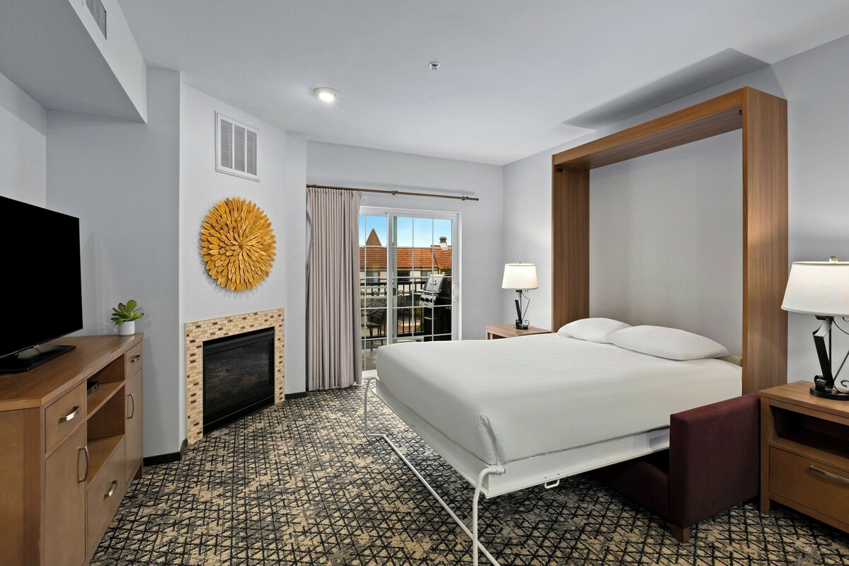 A hotel room with a bed, a television, a fireplace, a window with curtains, and a view of buildings outside.