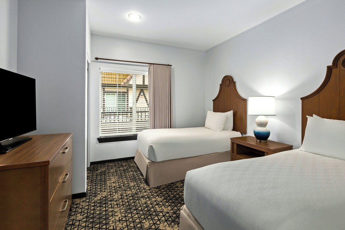 A hotel room with two beds, a TV, a lamp, and a window with blinds.