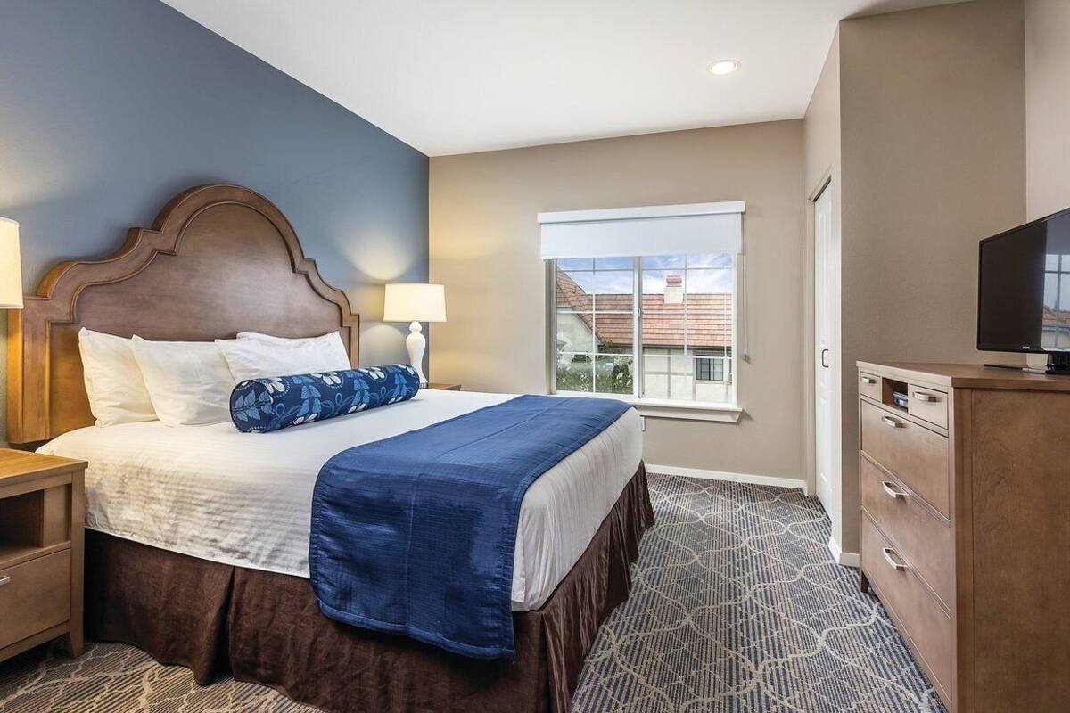 A hotel room with a large bed, two bedside tables with lamps, a dresser, a television, and a window with a view of buildings.