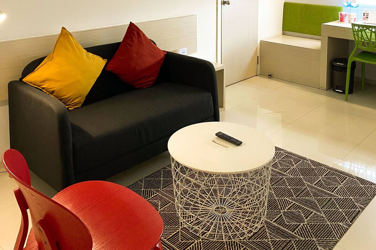 A modern living room with a black sofa, red and yellow cushions, a white coffee table, and a green chair. There is a framed picture on the wall and a small table with a remote control.