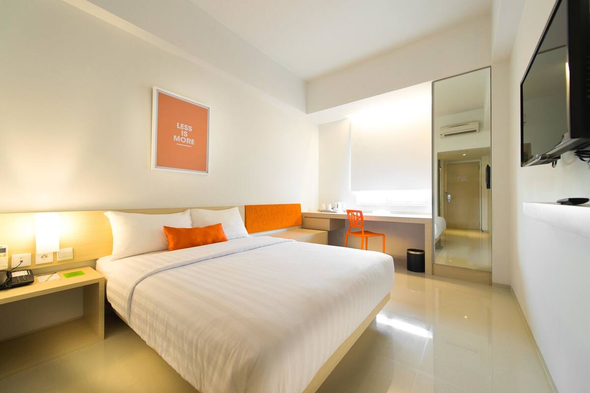 A modern hotel room with a large bed, a desk, and a chair. There is a bathroom with a glass door and a shower.
