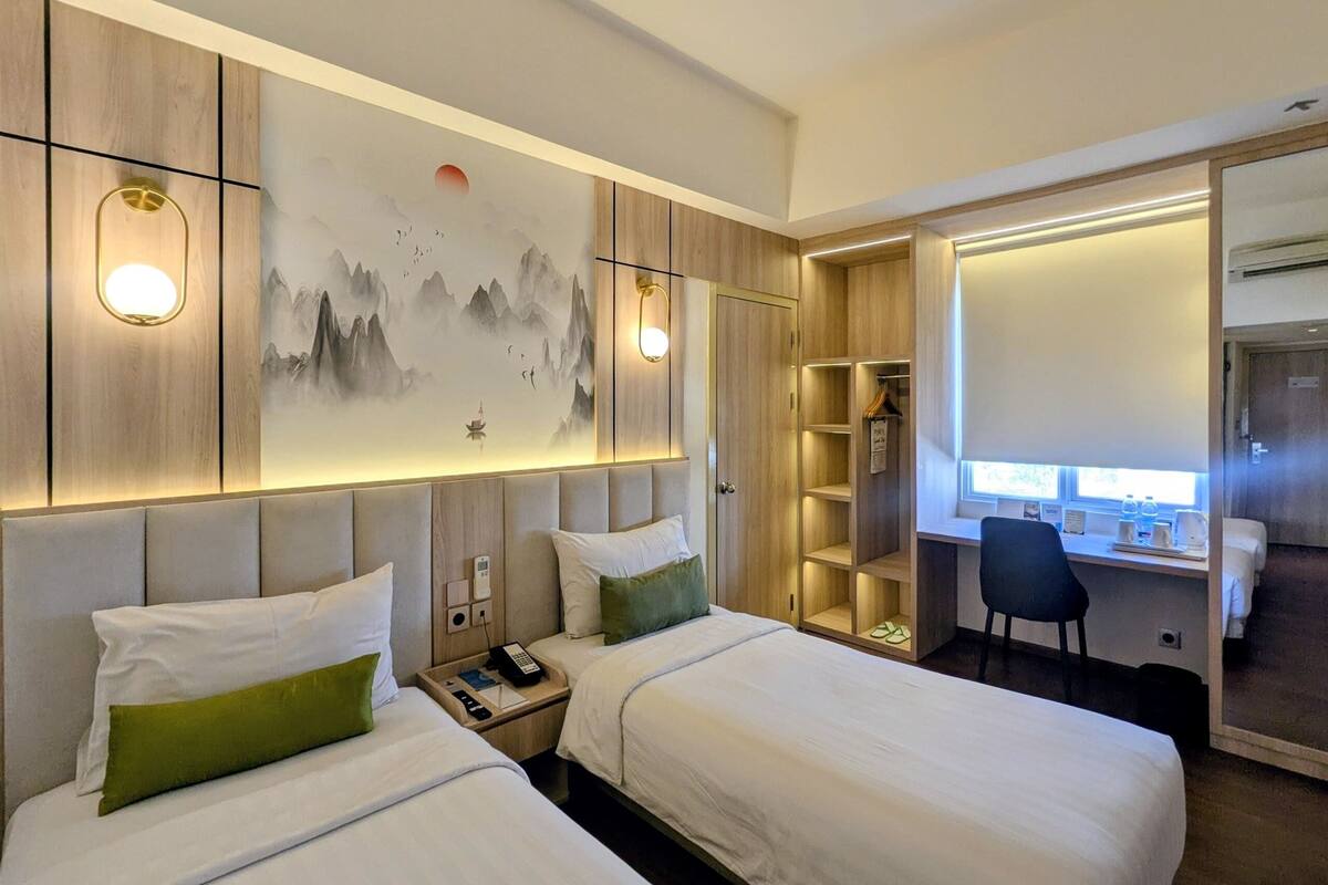 A hotel room with two beds, a desk, a chair, a wardrobe, and a painting on the wall.