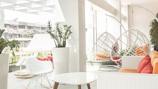 A modern indoor space with a white sofa, a round table, and hanging chairs. There are potted plants and a view of buildings outside. -  Ayda Mangga Dua d/h Zuri Express Mangga Dua (Jakarta)