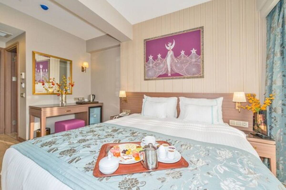 A hotel room with a bed, a desk, a mirror, a nightstand, and a painting on the wall.