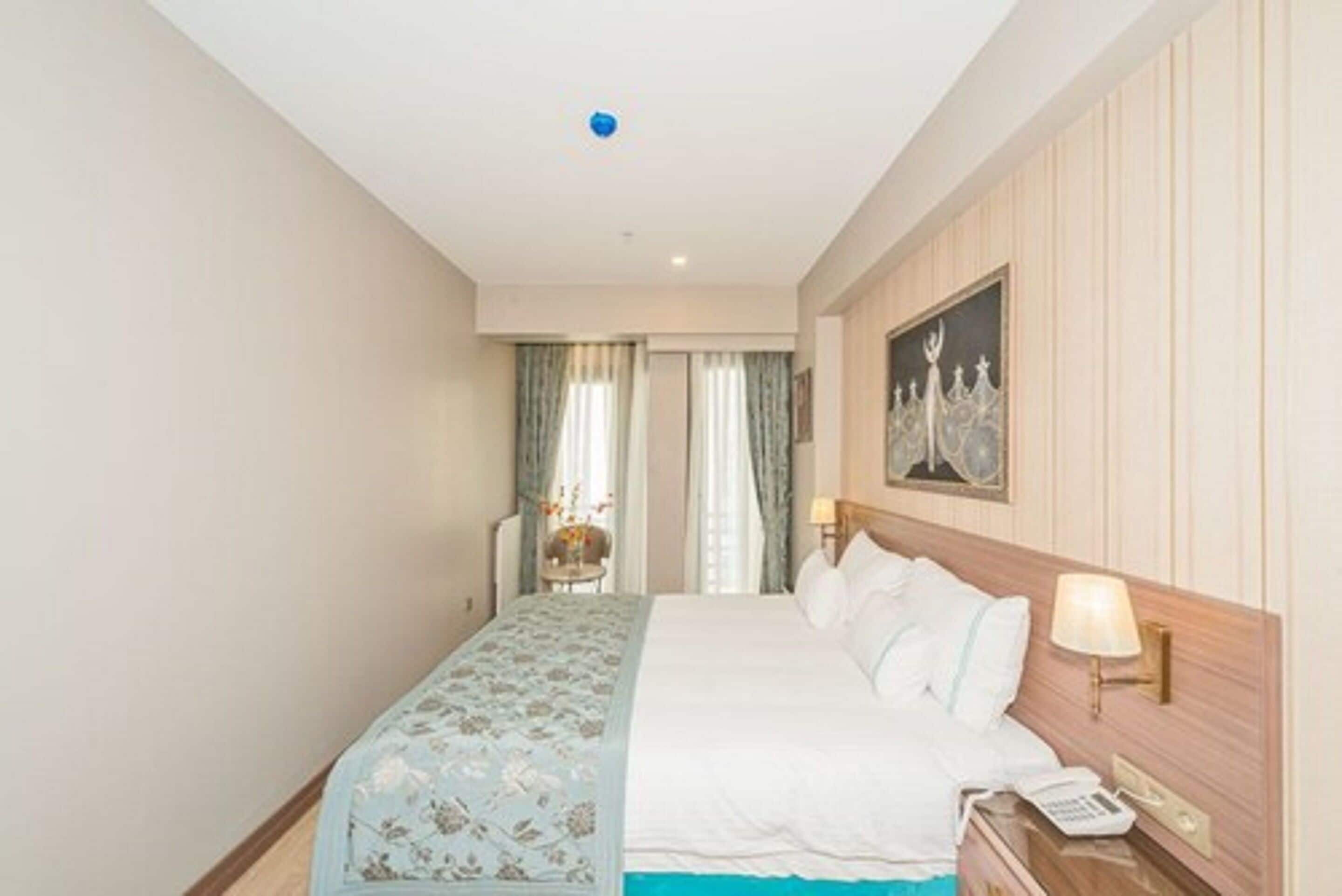 A hotel room with a bed, a bedside table with a lamp, a painting on the wall, and a view of the hallway.