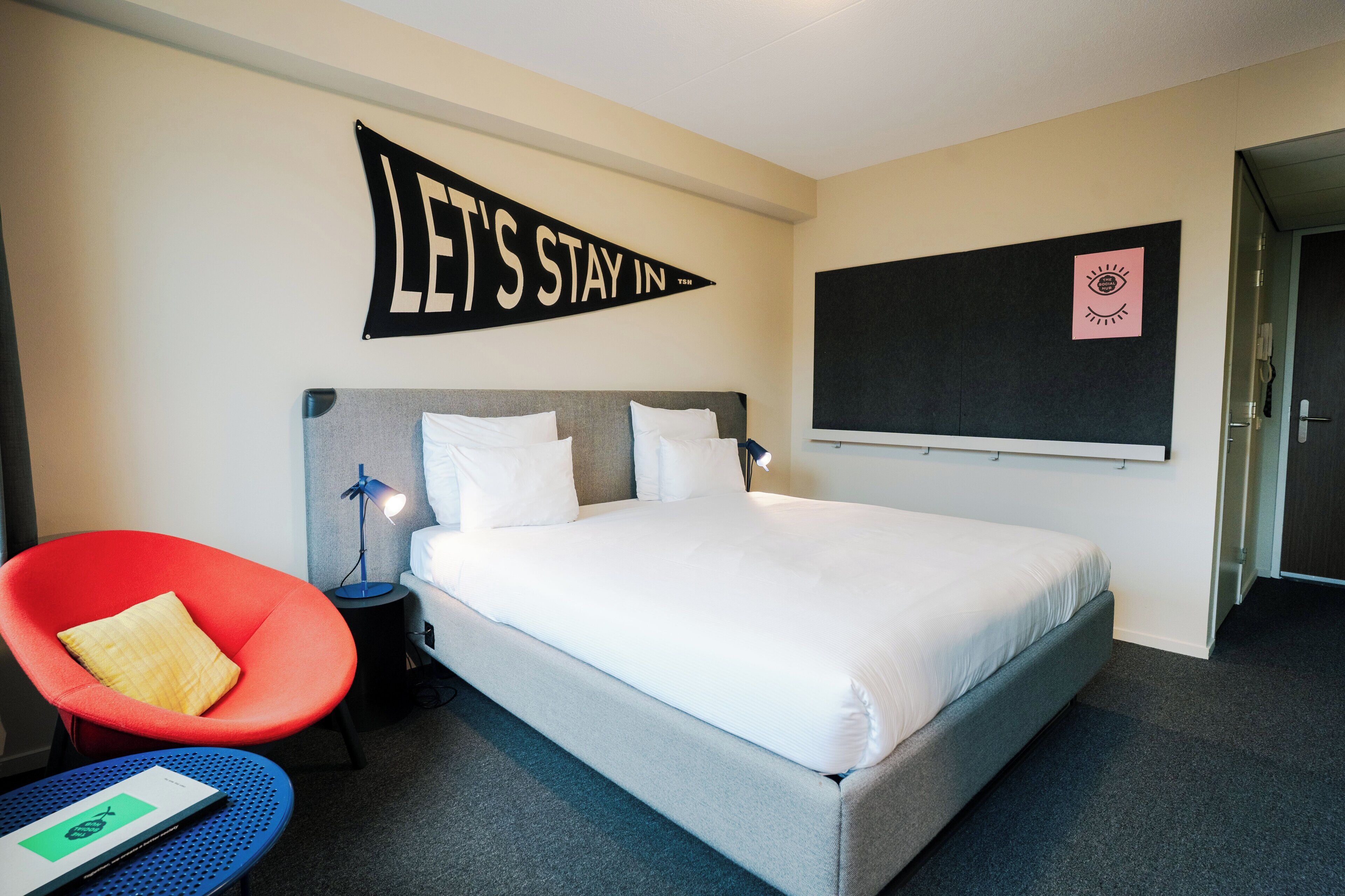 Ein Hotelzimmer mit einem großen Bett, einem roten Sessel, einem kleinen Tisch mit einem Buch und einem motivierenden Spruch an der Wand.