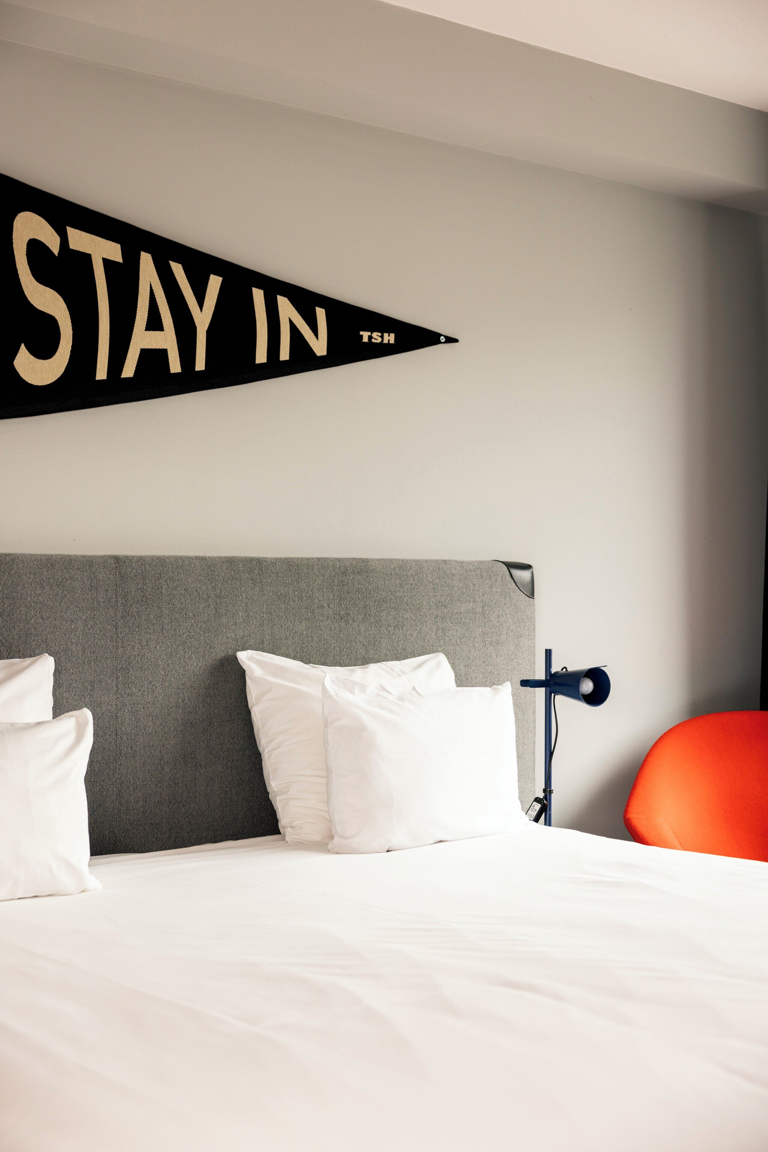 Ett hotellrum med en säng, vita kuddar, en grå sänggavel, en svart skylt med texten ”STAY IN” och en röd stol.