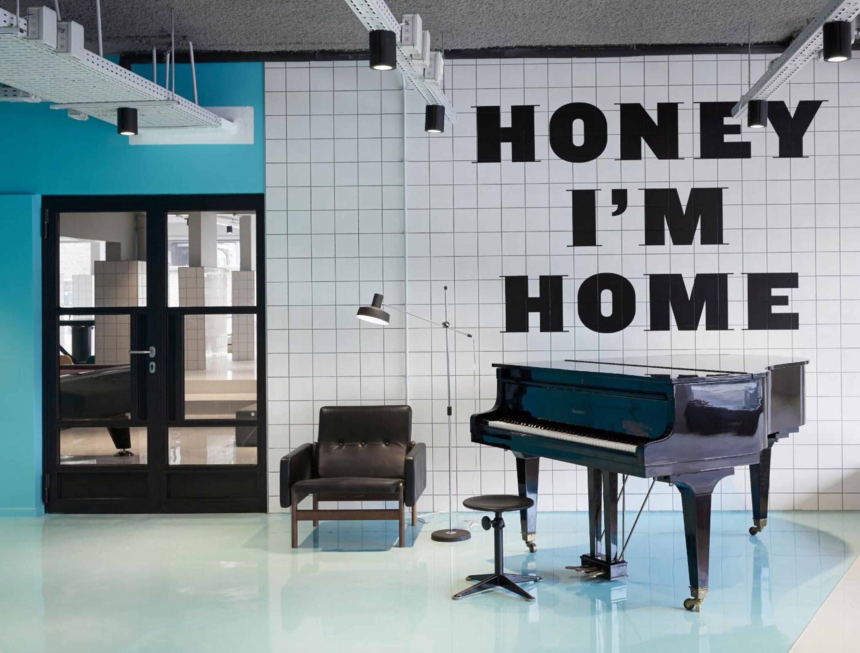 Huoneessa on piano, tuoli ja seinä, jossa lukee teksti: ”Rakas, olen kotona”.
