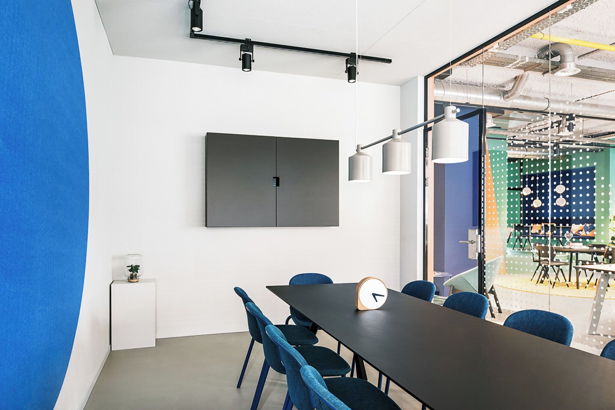 A conference room with a long table, blue chairs, a clock, and a TV on the wall.