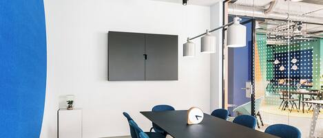 A conference room with a long table, blue chairs, a clock, and a TV on the wall.