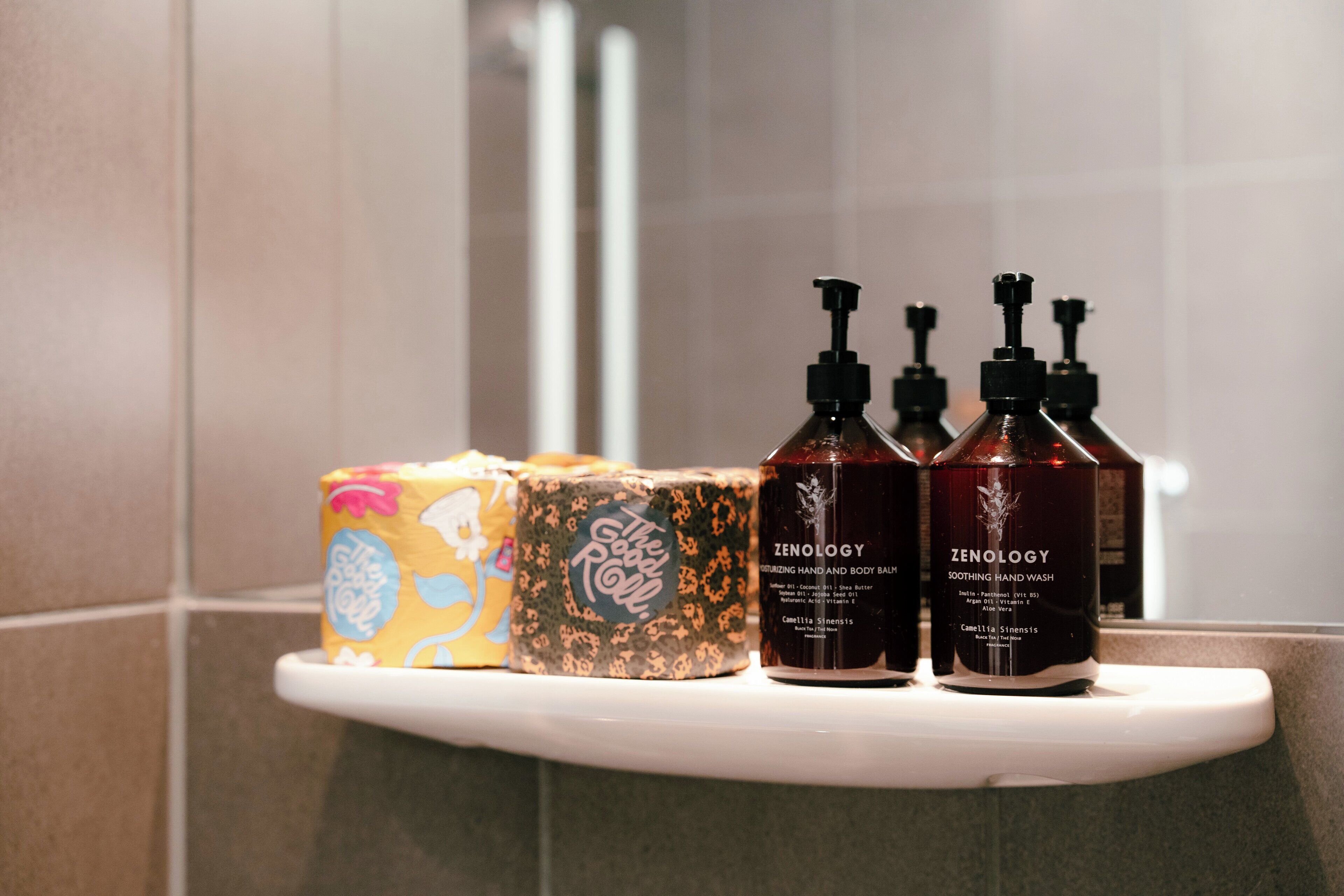 A bathroom shelf with toiletry items including two bottles of Zenology hand wash and two boxes of The Good Roll.
