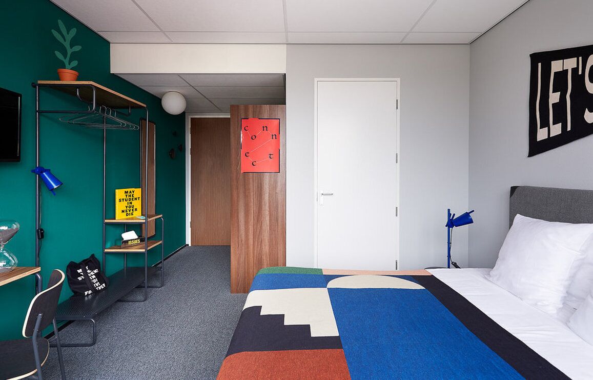 Une pièce comprenant un lit, un bureau, une chaise, une étagère avec un pot de plante, une horloge murale et une porte.