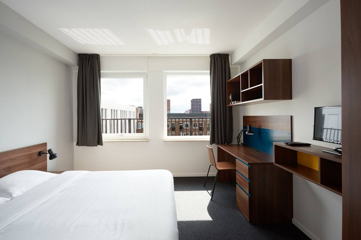 A hotel room with a bed, desk, chair, and a TV. There are windows with curtains and a view of buildings outside.