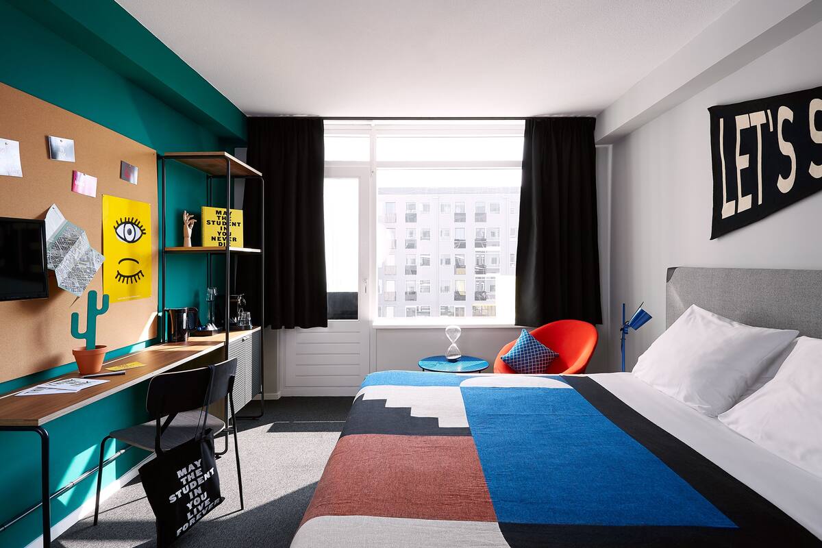 A modern hotel room with a bed, a desk, and a window with a city view.