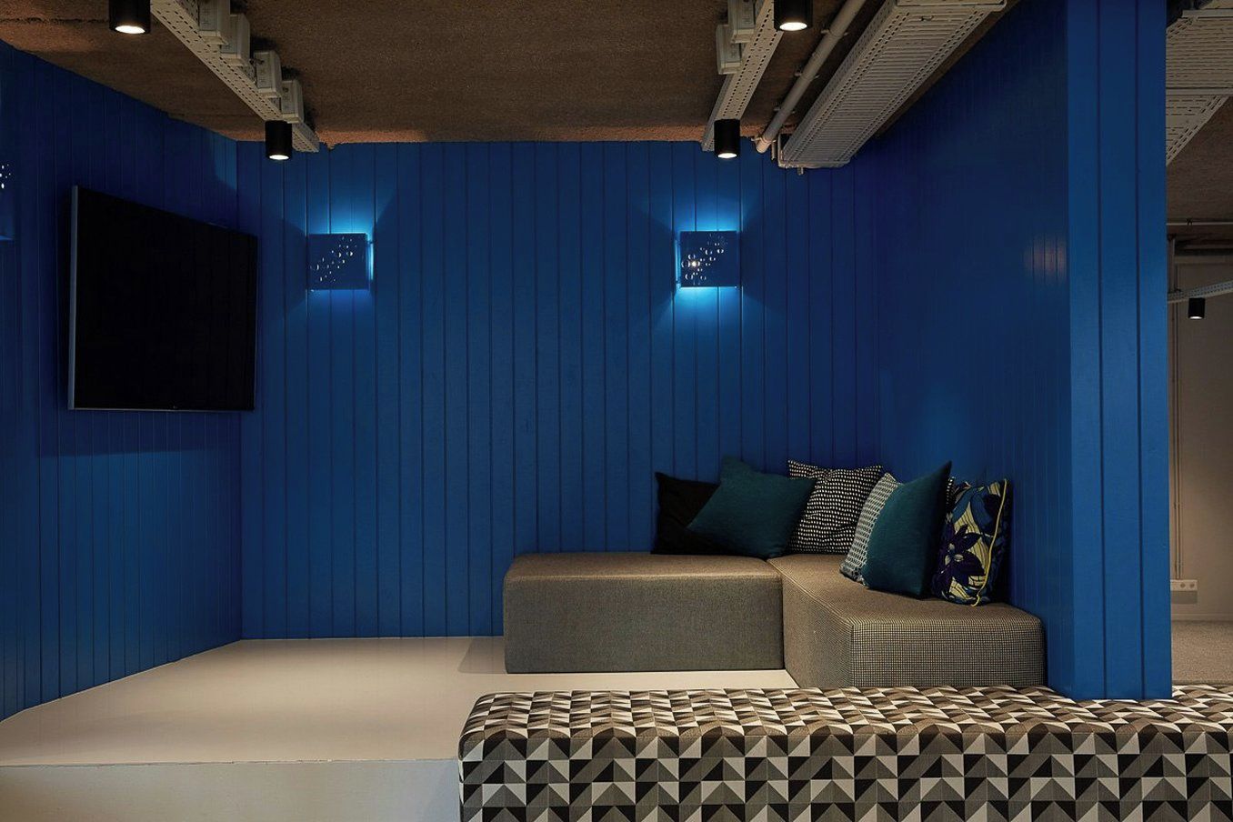 A room with blue walls, a flat-screen TV mounted on the wall, a sofa with cushions, and a patterned ottoman.