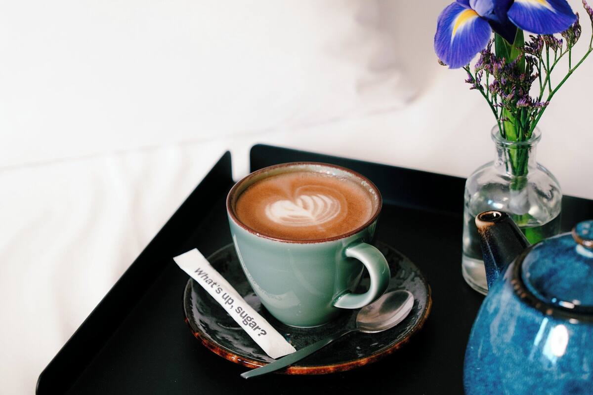 A tray with a cup of coffee, a small vase with flowers, and a teapot on a bed.