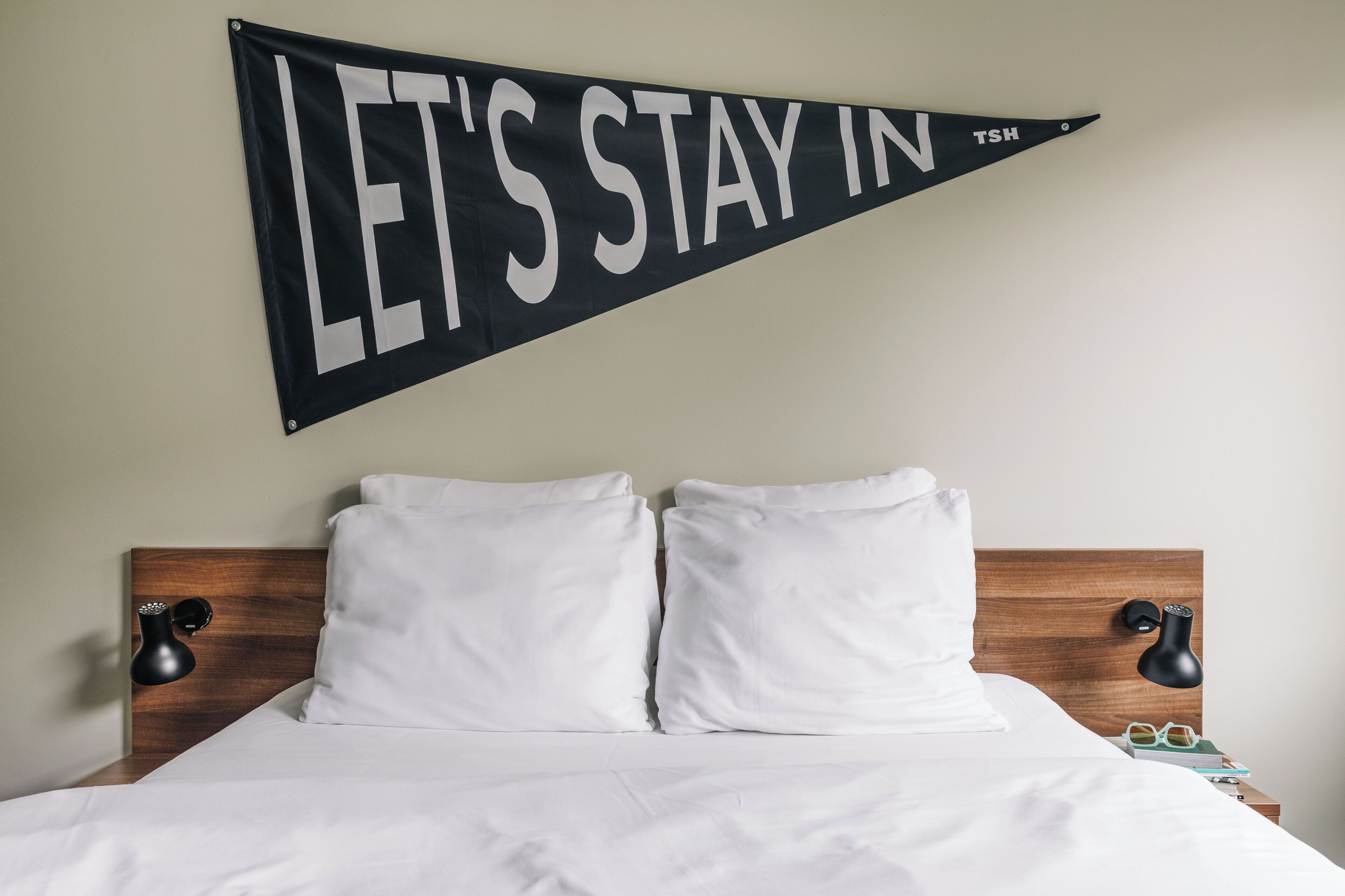 Un lit bien fait, avec des draps blancs, une tête de lit en bois et deux lampes de chevet. Une bannière noire avec du texte blanc est accrochée au-dessus du lit.