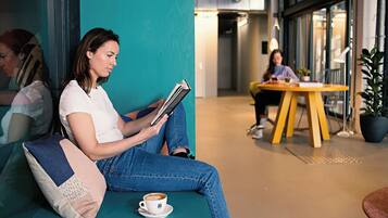 Una persona sentada en un sofá azul verdoso, leyendo un libro y tomando una taza de café, en un espacio interior moderno con una mesa amarilla. Se ve otra persona de fondo.