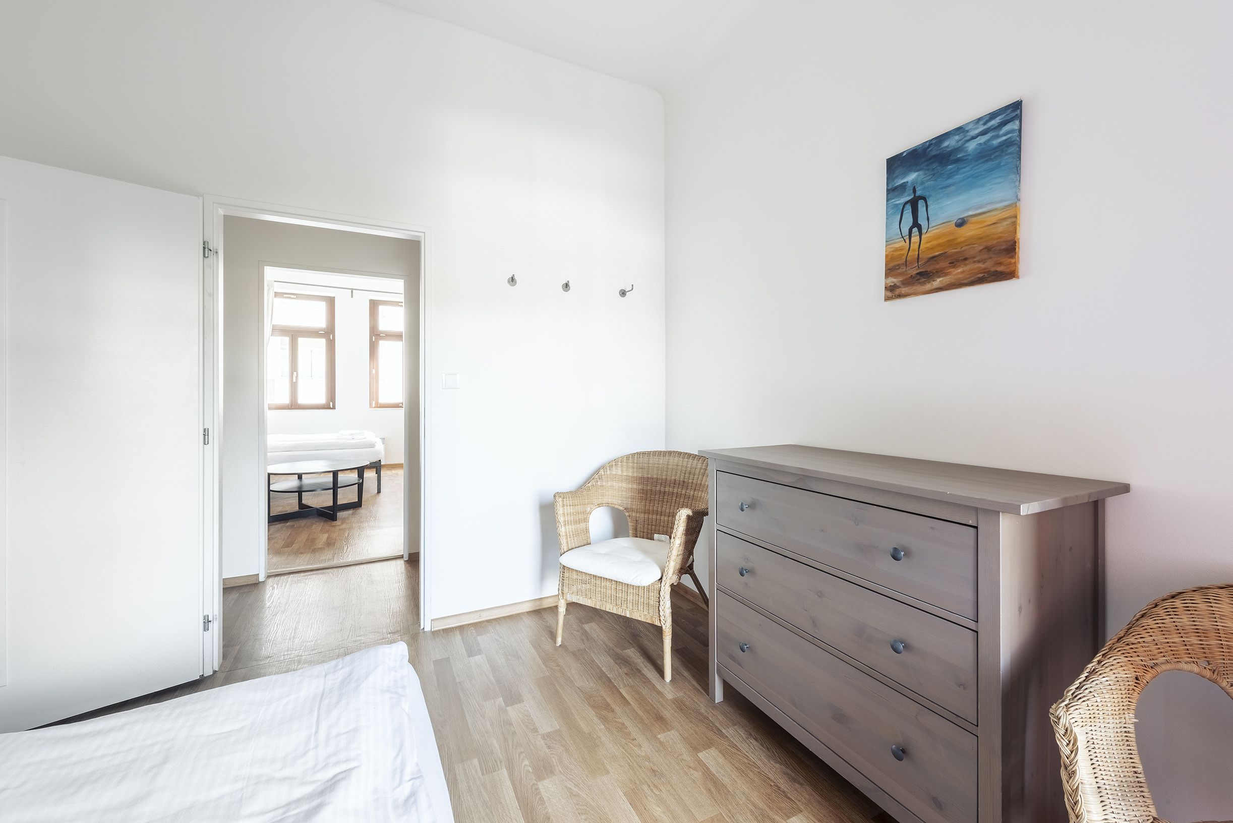 A bedroom with a bed, a dresser, a wicker chair, and a painting on the wall.