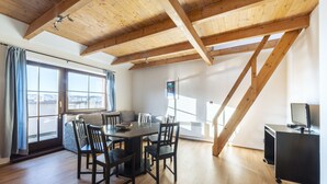 Une pièce spacieuse avec un plafond en bois, une table à manger avec des chaises, un téléviseur posé sur un meuble et de grandes fenêtres offrant une vue dégagée.