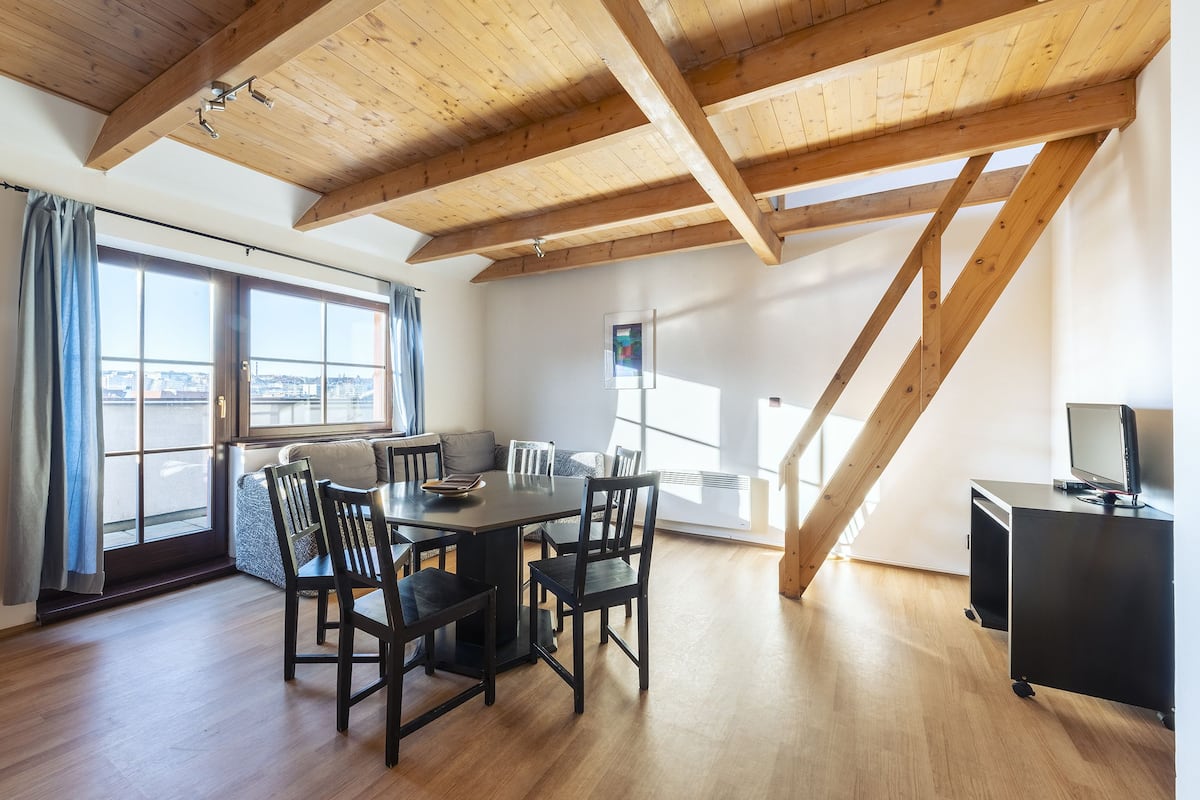 A spacious room with a wooden ceiling, a dining table with chairs, a TV on a stand, and large windows offering a view.