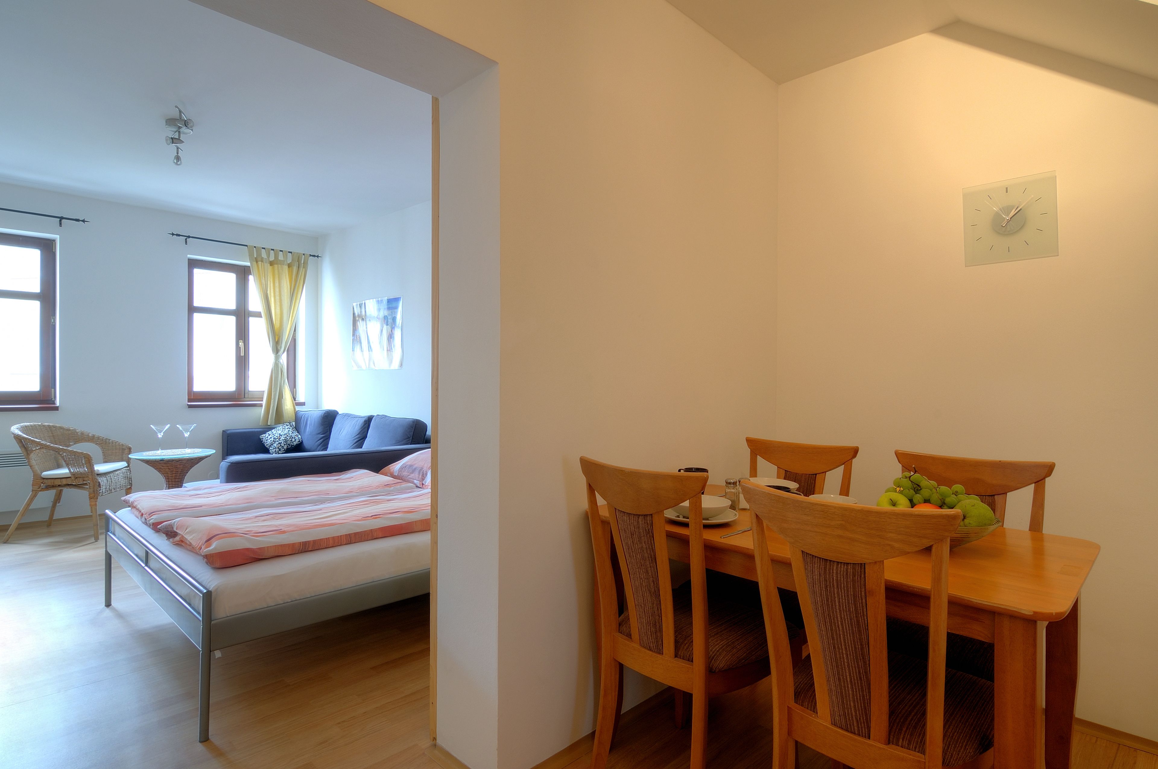 Un dormitorio con cama, mesita de noche y ventana con cortinas. Hay una zona de comedor con mesa, sillas y un bol de fruta.