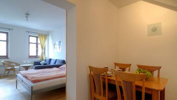 A bedroom with a bed, bedside table, and a window with curtains. There is a dining area with a table, chairs, and a bowl of fruit.