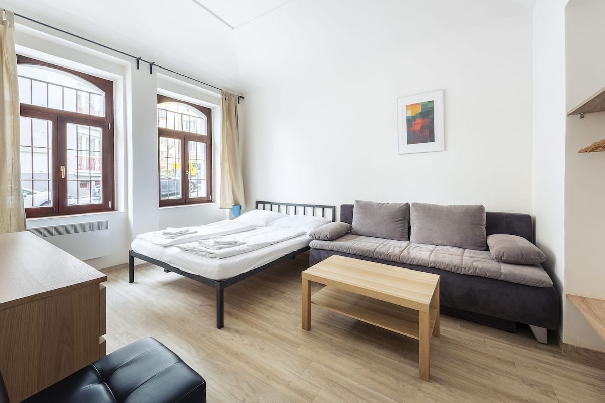 A modern, well-lit room with a bed, a sofa, a wooden table, and large windows with curtains.
