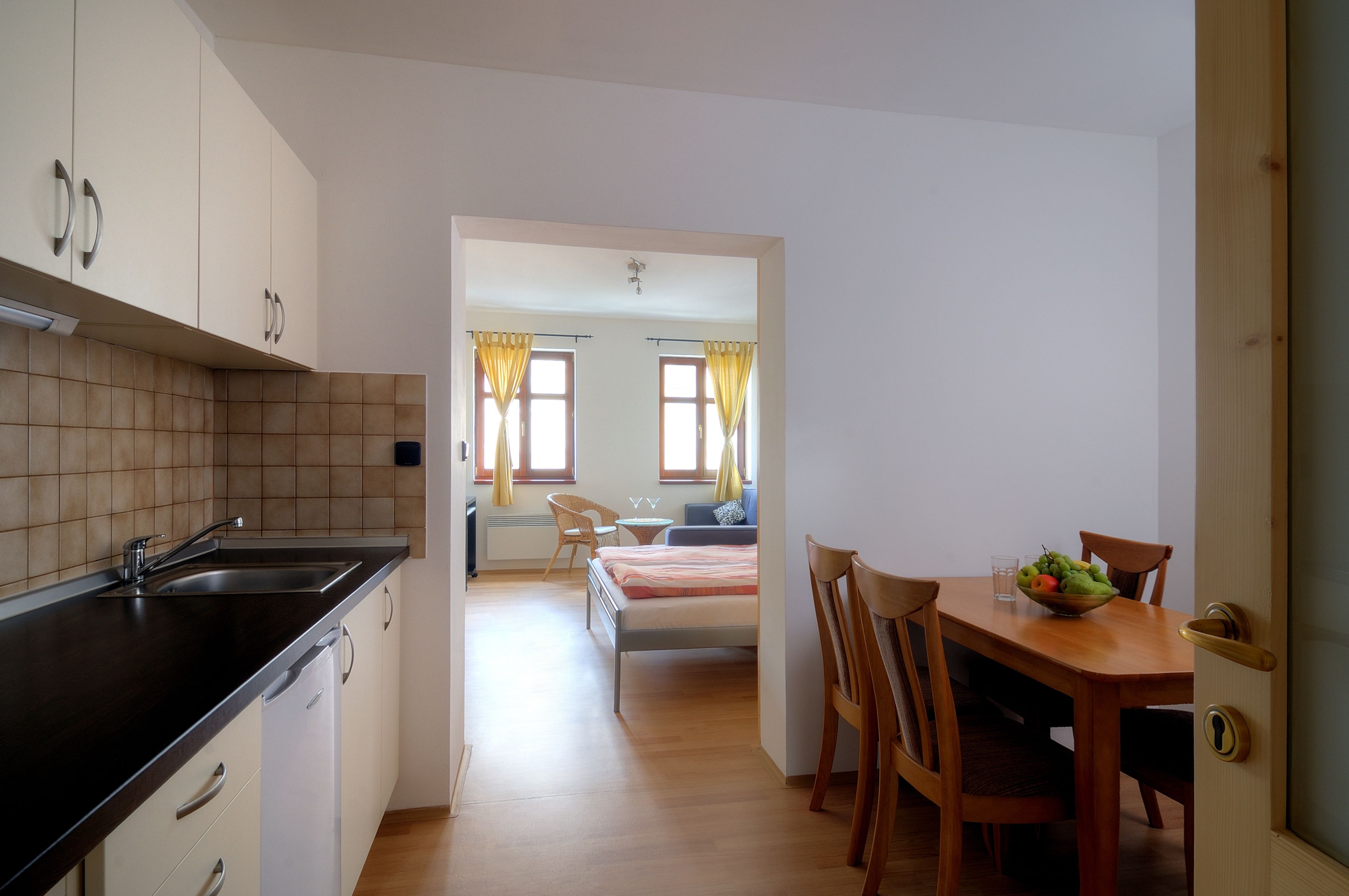 Een moderne keuken met een zwart aanrecht en witte kasten, die uitkomt op een eetgedeelte met een houten tafel en stoelen, en een slaapkamer met een bed en een raam met gordijnen.