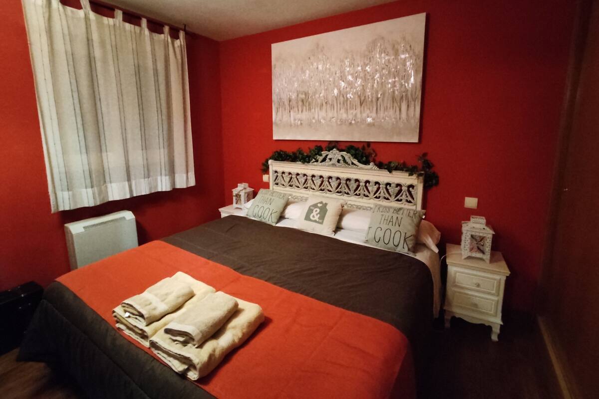 A hotel room with a red wall, a bed with a brown bedspread, a white nightstand, a white radiator, and a window with white curtains.