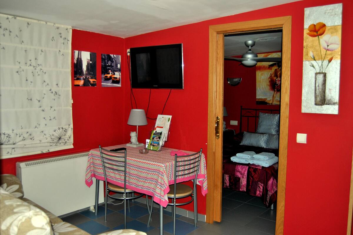 A dining area with a table set for two, a television mounted on the wall, and a door leading to another room.
