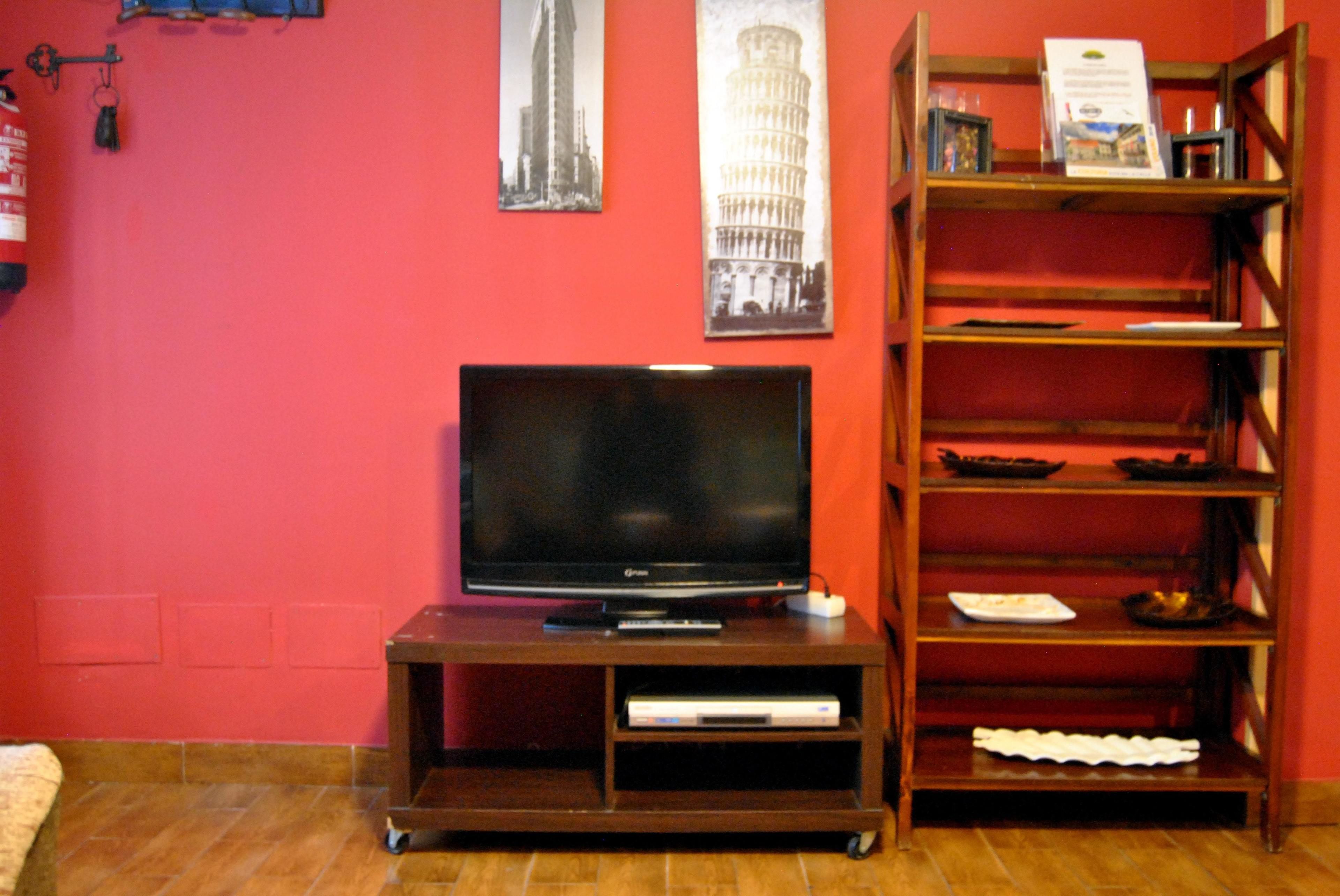 Ein Wohnzimmer mit einem Flachbildfernseher auf einem Holzständer, einem Bücherregal und einer roten Wand mit zwei gerahmten Bildern.