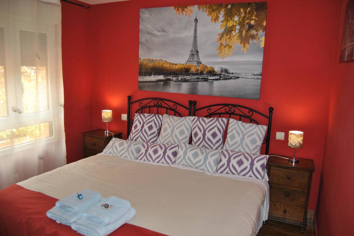 A bed with patterned pillows, a red wall, a painting of the Eiffel Tower, and two bedside lamps.