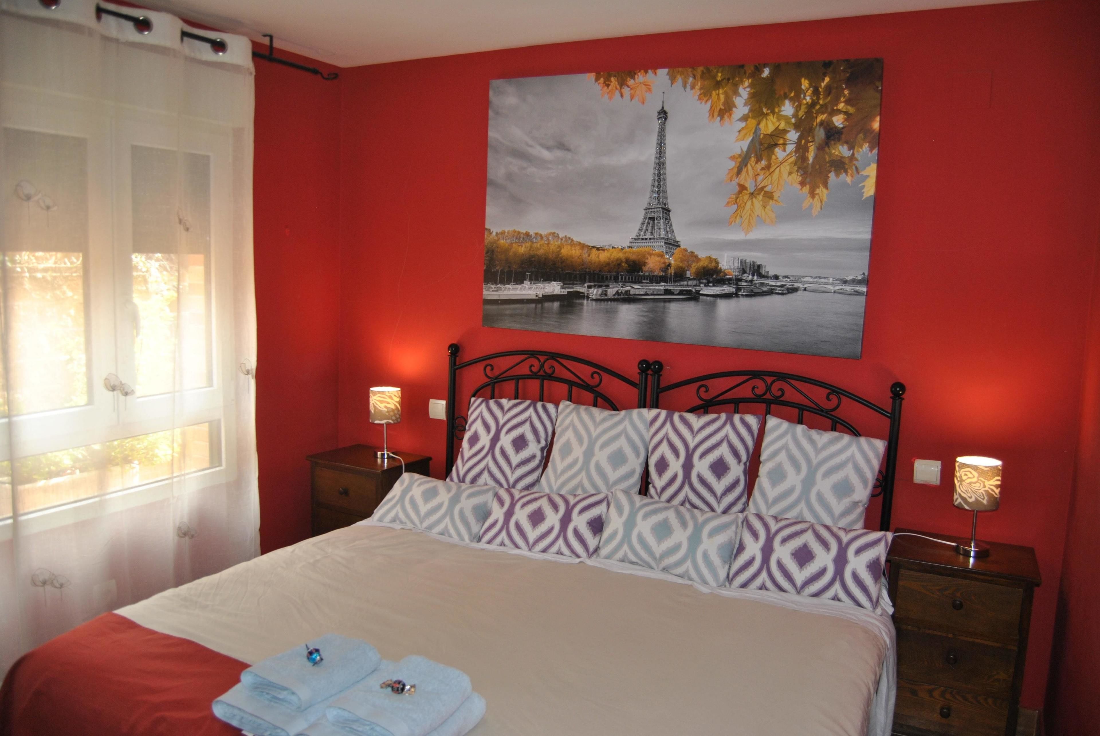 A bedroom with a bed, two bedside tables, a window with curtains, and a wall-mounted picture of the Eiffel Tower.