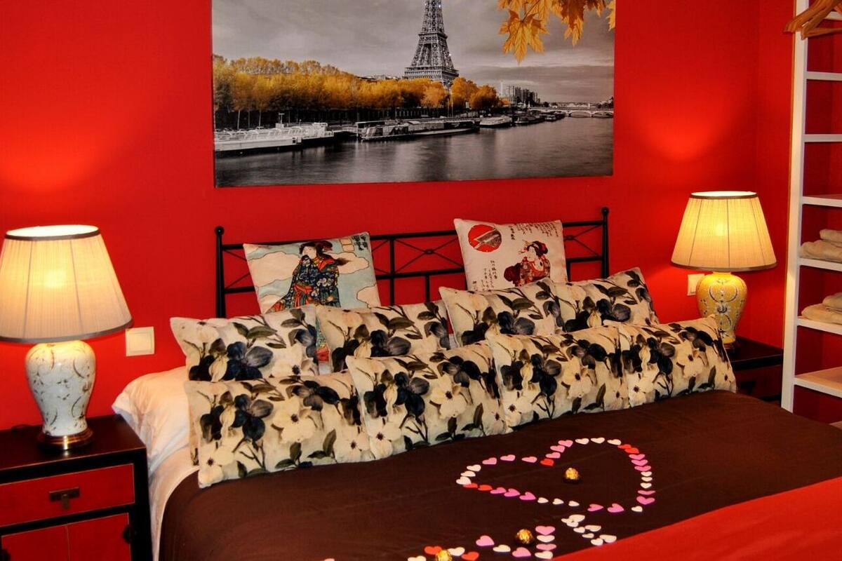 A bedroom with a red wall, a bed with floral pillows, a heart-shaped decoration on the bed, and a painting of the Eiffel Tower.