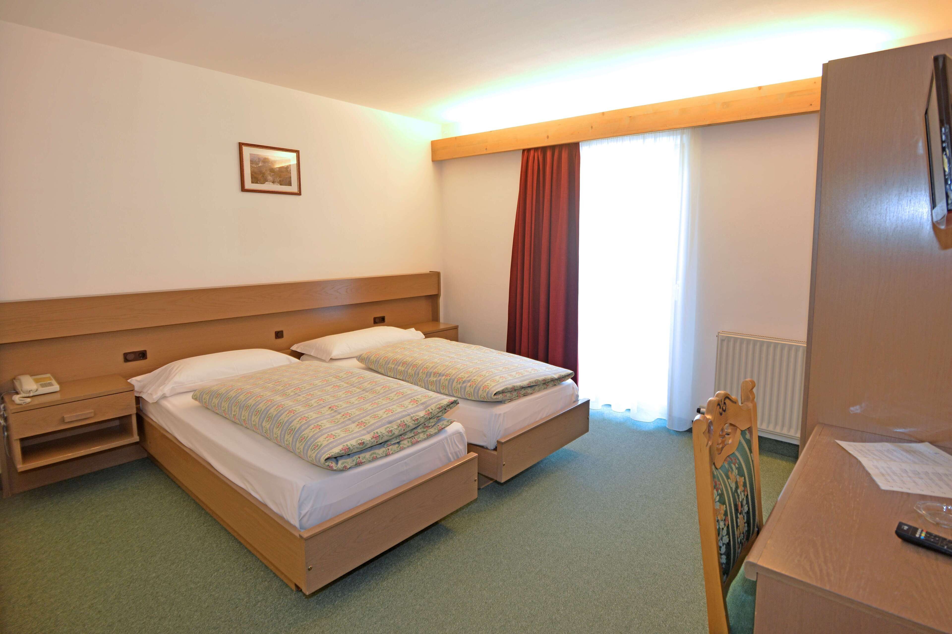A hotel room with two single beds, a wooden headboard, a small bedside table with a phone, a chair, a desk with a remote control, and a window with red curtains.