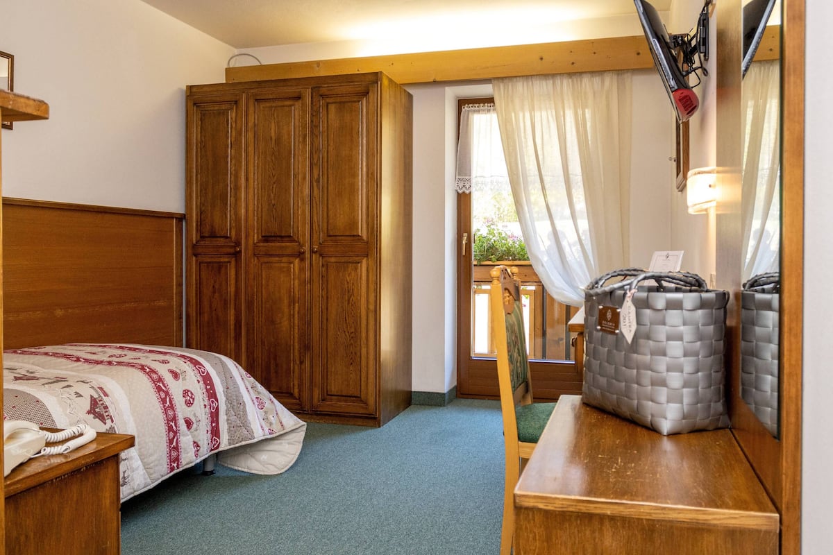 Une chambre à coucher avec un lit en bois, une armoire, un bureau avec une lampe et une vue sur l’extérieur par une fenêtre ornée de rideaux.