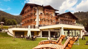A multi-story building with balconies, outdoor seating, and a grassy area with stacked logs. - Wellness Hotel Dolomia (Soraga)