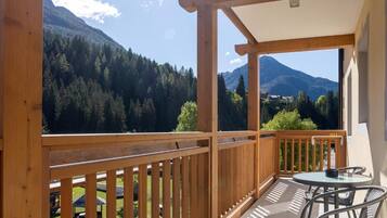 Un balcone con ringhiera in legno, un tavolo con sedie e vista su una catena montuosa ricoperta di boschi.