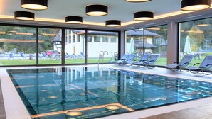 An indoor swimming pool with clear glass sides, surrounded by lounge chairs and large windows offering a view of the outside. - Wellness Hotel Dolomia (Soraga)