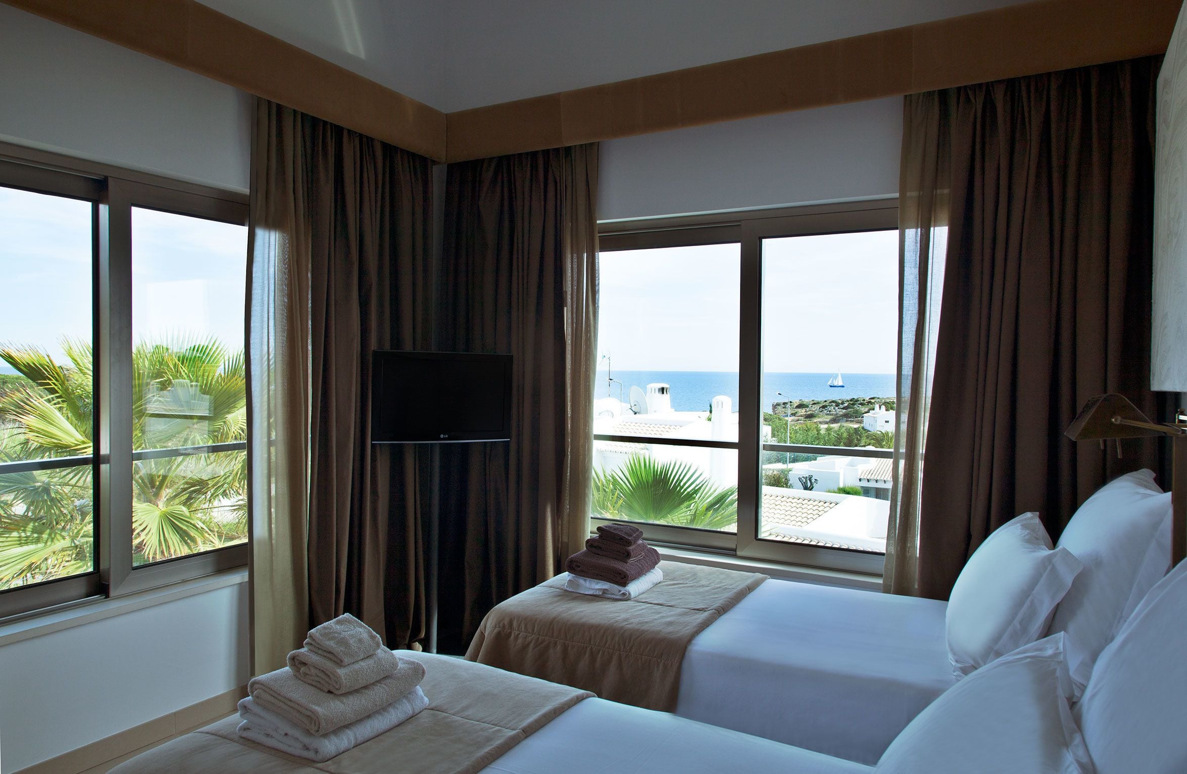 Une chambre d’hôtel avec deux lits, une télévision et de grandes fenêtres donnant sur la mer et les palmiers.