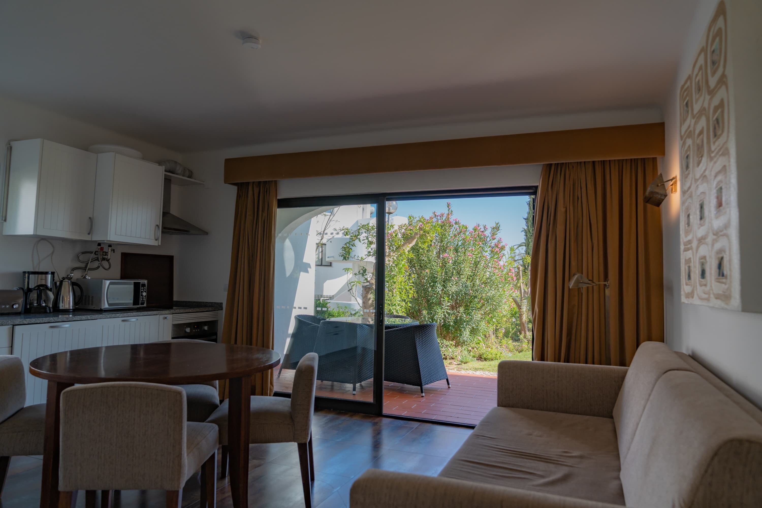 A modern living room with a sofa, a dining table, and chairs. There is a kitchen area with cabinets and a microwave. The room has a sliding glass door leading to an outdoor area with a table and chairs.
