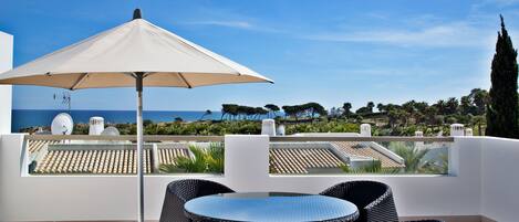 A terrace with a table and chairs, a large umbrella, and a view of the sea and trees.