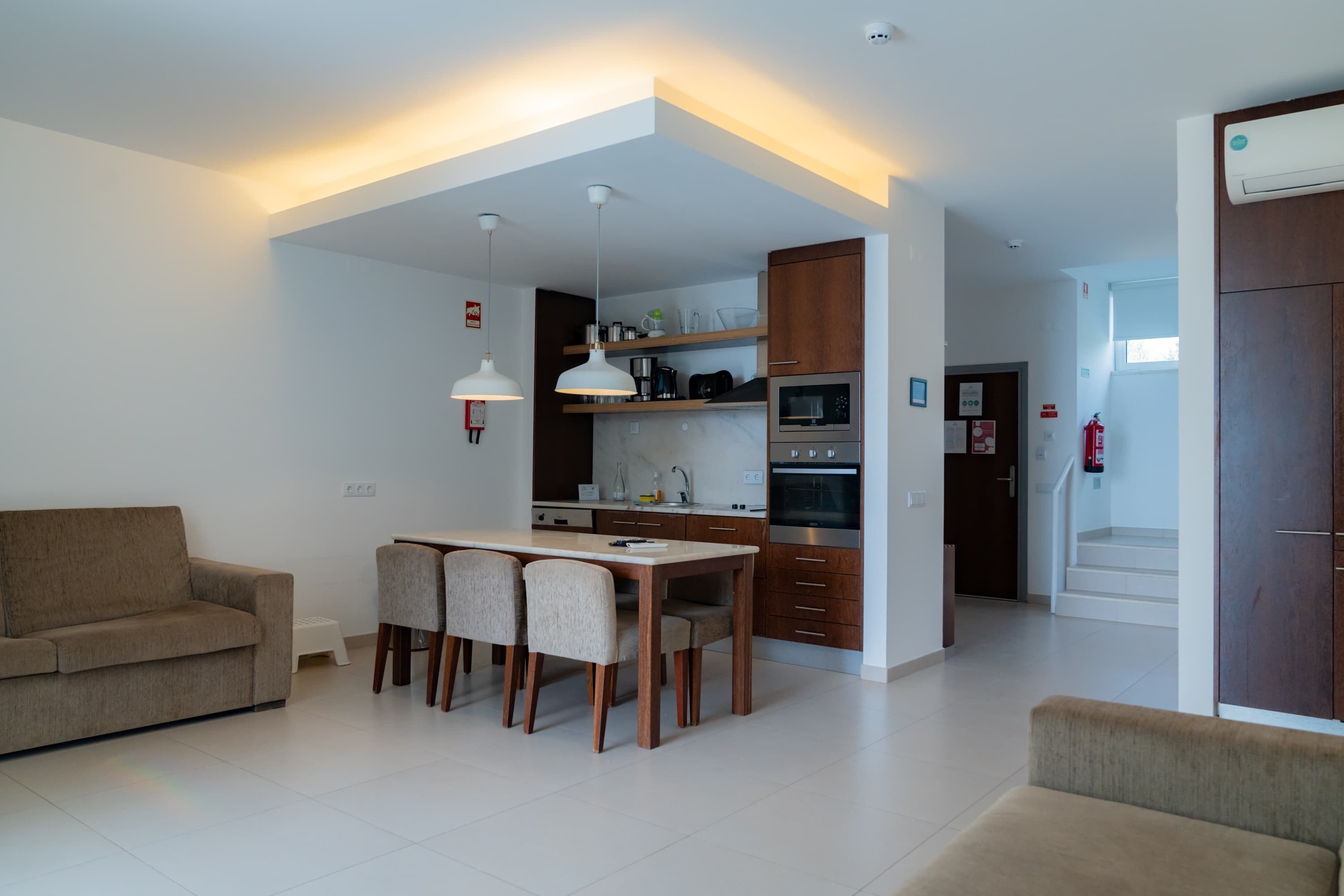 Una sala de estar moderna con un sofá, mesa de comedor y sillas. Cuenta con una zona de cocina con electrodomésticos y una escalera que lleva a otro nivel.