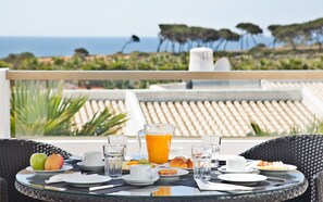 A breakfast table with plates, cups, and a pitcher of orange juice, overlooking a scenic view of the sea and trees.