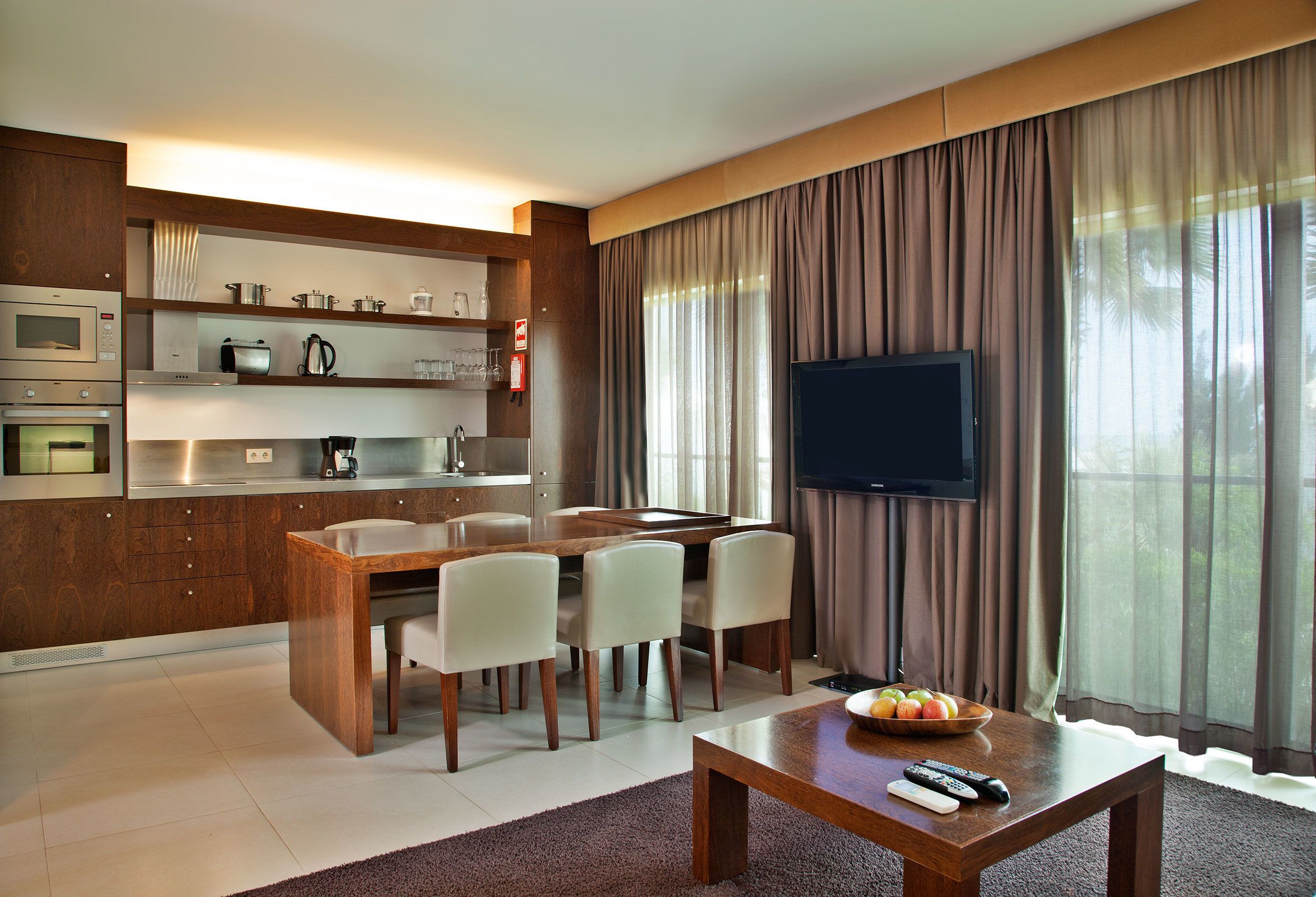Una cocina moderna con electrodomésticos integrados, un comedor con mesa y sillas de madera, un televisor de pantalla plana y un frutero sobre una mesita de centro.