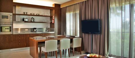 A modern kitchen with built-in appliances, a dining area with a wooden table and chairs, a flat-screen TV, and a fruit bowl on a coffee table.