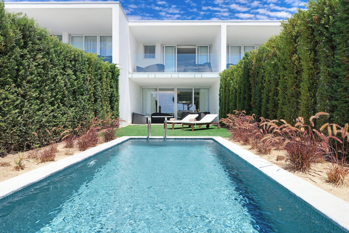Una zona de piscina moderna amb una piscina de blau clar, gandules i un edifici de dues plantes amb grans finestres.